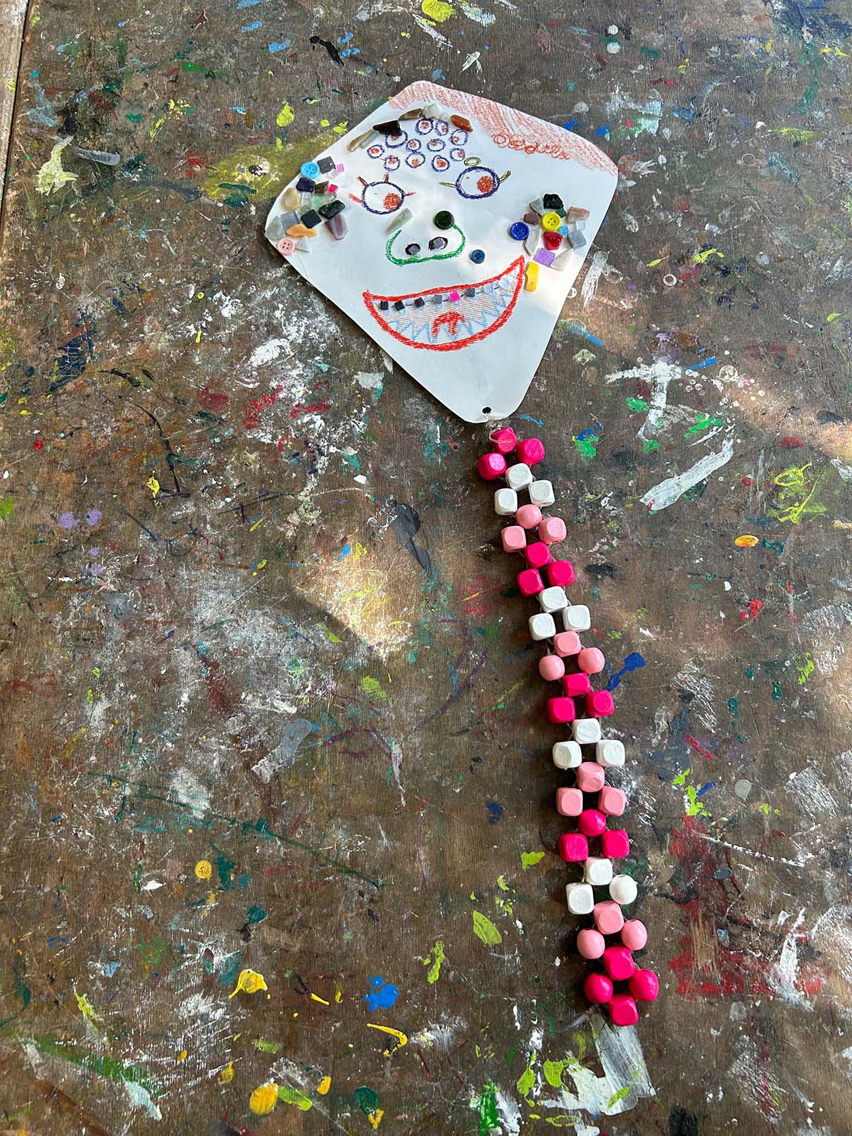 Ein selbstgemachter Flugdrache mit Gesicht liegt auf einem dunklen Untergrund mit Farbkleksen