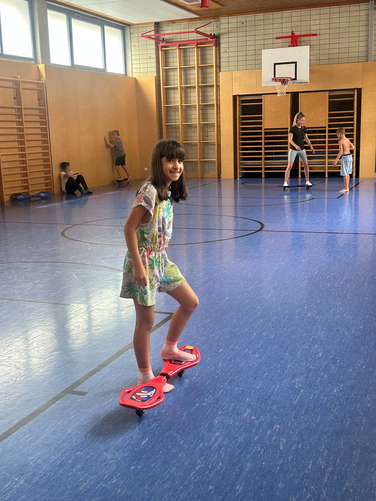 Ein Mädchen steht mit einem Bein auf einem roten Waveboard und lächelt in die Kamera