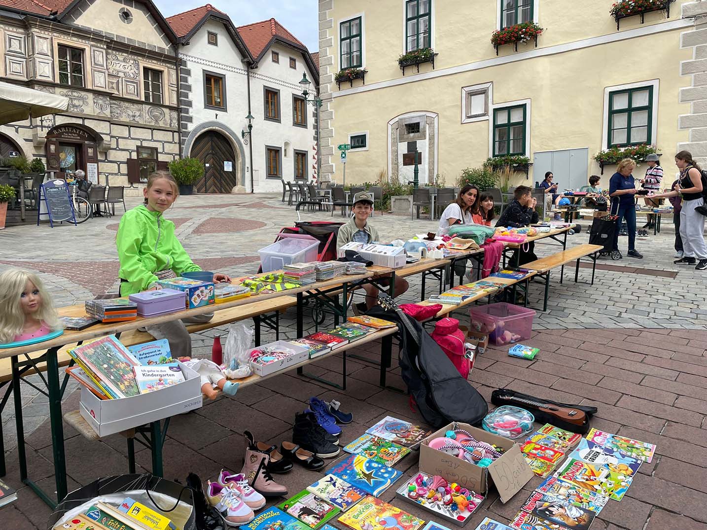 Mehrere Kinder sitzen verteilt bei einem langen Tisch voll mit Spielzeug, Büchern und Boxen