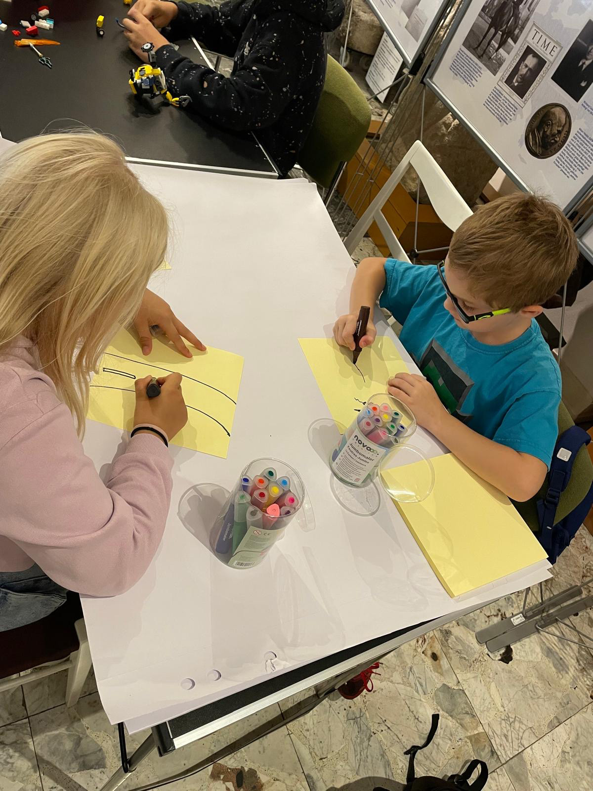 Zwei Kinder sitzen bei einem Tisch und zeichnen etwas auf Papier