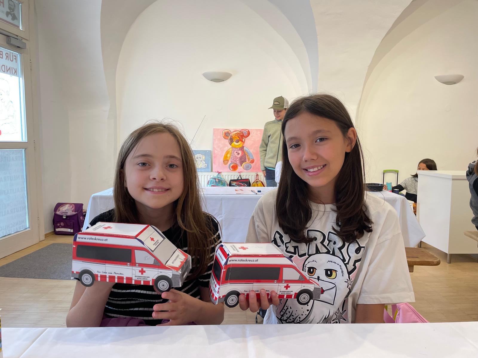 Zwei Mädchen lächeln und schauen in die Kamera und zeigen die selbstgebauten Papprettungswagen her.