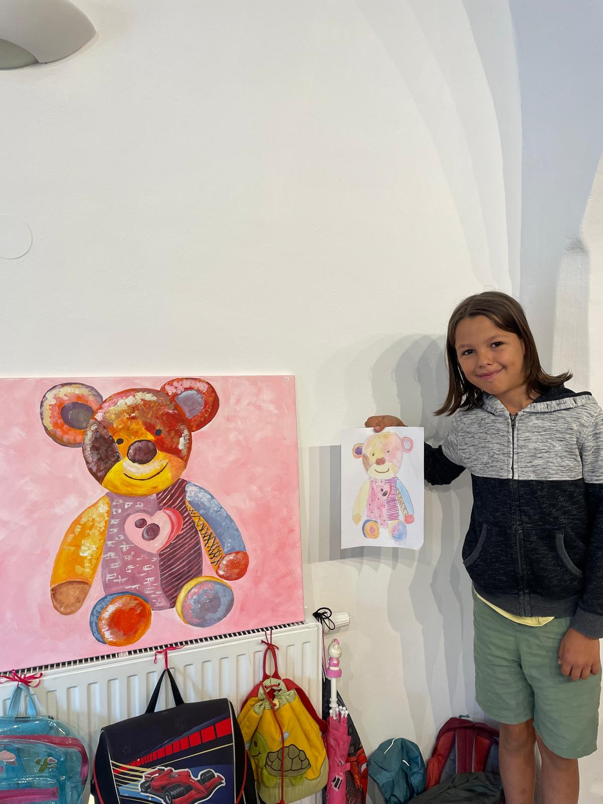 Ein Mädchen steht neben einem Bild mit einem Stoffbären und zeigt ein selbstgemaltes Bild von dem Stoffbären her.